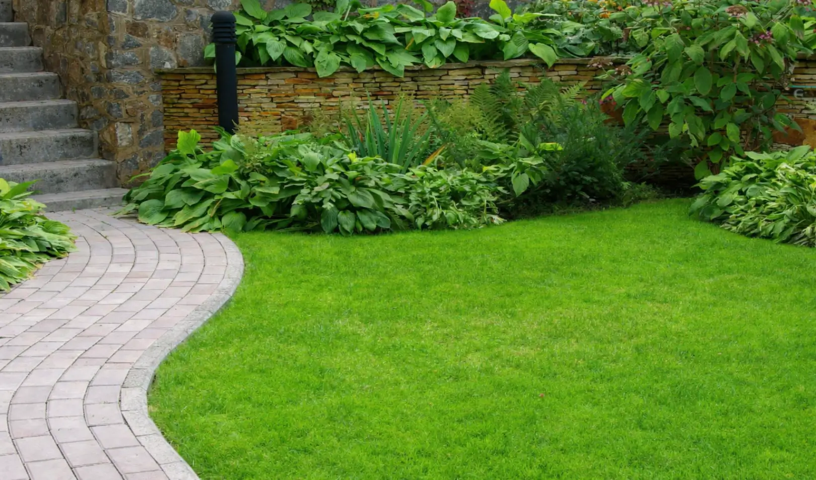 Uređena okućnica sa zelenim travnjakom, zakrivljenom popločenom stazom, kamenim stepenicama i bogatim zelenim raslinjem uz kameni zid.